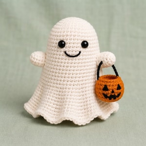Puede incluir: Una figura de fantasma tejida a ganchillo de color crema con cabeza redondeada y base acampanada. El fantasma tiene dos pequeños ojos de cuentas negras y una sonrisa negra bordada. Sostiene un pequeño cubo de calabaza naranja con asa negra.