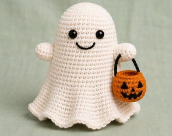 May include: A cream-colored crocheted ghost figurine with a rounded head and flared base. The ghost features two small black bead eyes and a stitched black smile. It holds a small orange crocheted pumpkin bucket with a black handle and face.