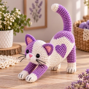 May include: A crocheted cat toy featuring a white and purple color scheme. The cat is in a stretching pose with a purple heart-shaped patch on its back, a pink nose, black plastic eyes, and black thread whiskers. The tail is held upright.