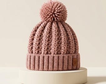 May include: A dusty rose knit beanie featuring a vertical ribbed texture and a large matching pom-pom on top. The hat includes a wide folded brim with a small square brown leather patch on the side, which is embossed with a simple heart outline.