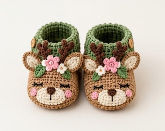 May include: A pair of handmade crochet baby booties designed to look like deer. The booties feature a tan base with a green ribbed cuff, brown antlers, pink and white floral accents, and embroidered closed eyes with pink cheeks.