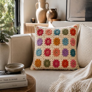 Puede incluir: Un cojín cuadrado con una funda de ganchillo de color crema que presenta una cuadrícula de dieciséis motivos florales individuales. Cada flor está realizada en un color vibrante diferente, incluyendo tonos de rojo, naranja, amarillo, verde, azul y morado.
