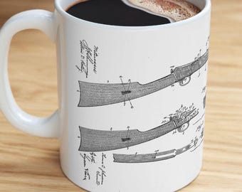 May include: White ceramic coffee mug featuring a detailed black and white diagram of a rifle. The design includes technical drawings and handwritten text. The mug is filled with dark coffee and has a white handle.