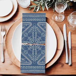 May include: A folded blue and cream-colored napkin with an intricate pattern, tied with twine, rests on a white plate. The napkin features a symmetrical design with floral and geometric elements. The setting includes a wooden charger, silverware, and glassware.