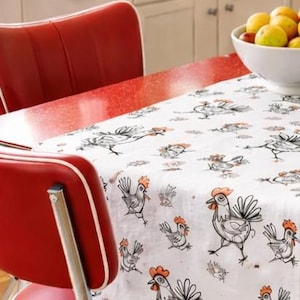 May include: A red retro-style chair and a table covered with a white tablecloth featuring a black and orange chicken pattern. A bowl of fruit sits on the table. The table has a red countertop.