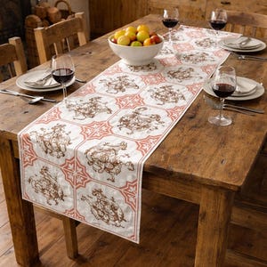 Puede incluir: Una mesa de comedor rústica de madera preparada para una comida. Un camino de mesa blanco y melocotón con ilustraciones marrones de figuras está centrado en la mesa. Copas de vino, platos y un cuenco de fruta completan el conjunto.