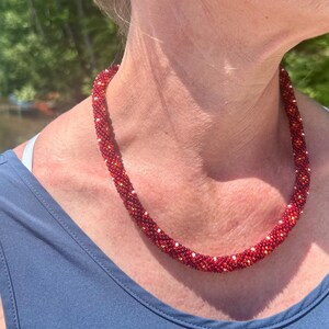 May include: A tubular beaded necklace featuring a dense, spiraling pattern of small, deep red beads interspersed with tiny, reflective silver-toned accents. The necklace has a uniform, cylindrical shape that rests comfortably around the neck.