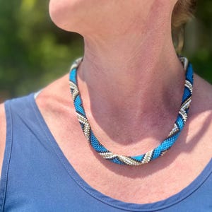 May include: A tubular beaded necklace featuring a spiral pattern of bright turquoise, dark navy blue, and metallic silver beads. The necklace is constructed from small, uniform beads arranged in a continuous, flexible rope design that rests around the neck.