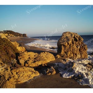 Puede incluir: Un paisaje costero que muestra una playa de arena con grandes formaciones rocosas de color marrón dorado en primer plano. Las olas blancas del océano rompen en la orilla bajo un cielo azul claro, con un acantilado visible a lo lejos.