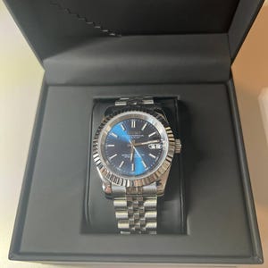 Puede incluir: Un reloj de pulsera plateado con esfera azul, bisel estriado y brazalete de eslabones metálicos, presentado en una caja negra con el nombre de la marca SEIKO impreso en blanco en la tapa interior. El reloj cuenta con una ventana de fecha.