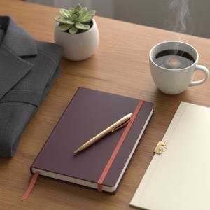 May include: A dark purple notebook with an orange elastic closure and matching ribbon bookmark, featuring a gold ballpoint pen resting on top. Beside it are a white ceramic mug of steaming coffee, a small succulent in a white pot, and a grey blazer.