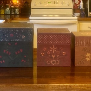 May include: A set of seven square wooden boxes arranged in descending order of size. Each box features a distinct color, including purple, dark blue, maroon, brown, and black, and is decorated with hand-painted floral and geometric patterns.