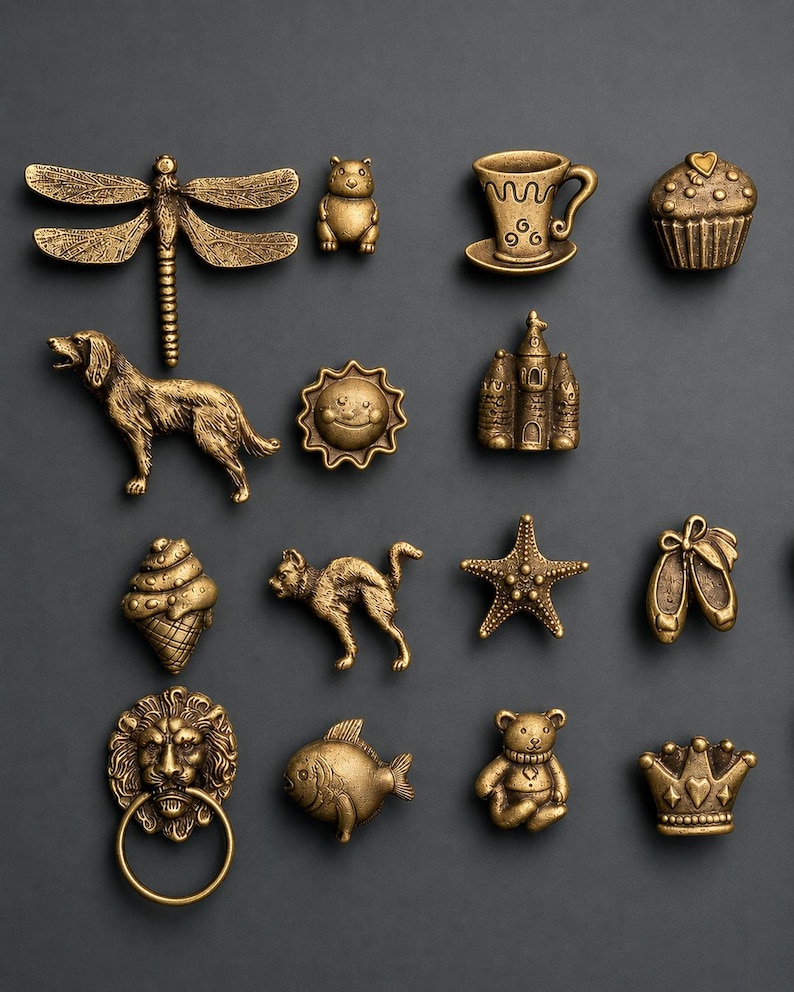 Pu&ograve; includere: Una collezione di pomelli per mobili in metallo color ottone antico con varie forme, tra cui una libellula, un cane, un gatto, una testa di leone con anello, un cupcake, una tazzina, un castello, un sole, una stella marina, scarpette da danza, un pesce, un orsetto e una corona.