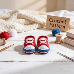 May include: A pair of handmade crochet baby sneakers in red, white, and blue. The shoes feature white laces, a white toe cap, and a blue sole trim. Beside the shoes are yarn balls, a wooden spool, crochet hooks, and a sign reading Crochet Pattern.