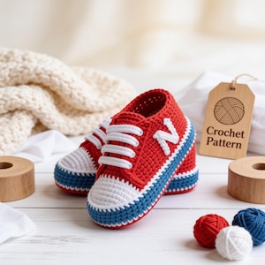 May include: A pair of red, white, and blue crochet baby shoes with white laces and a white letter N on the side. A wooden tag with the text Crochet Pattern is visible, along with small balls of red, white, and blue yarn on a white surface.