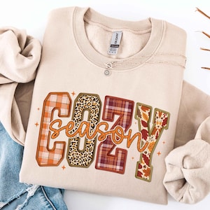 May include: A beige crewneck sweatshirt featuring the word COZY in large block letters with varied patterns including orange plaid, leopard print, maroon plaid, and autumn leaves, with the word season in orange cursive script overlaid.