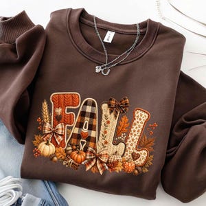May include: A dark brown crewneck sweatshirt featuring the word FALL in large, decorative letters. Each letter has a unique pattern including cable knit, plaid, and floral prints. The design is accented with pumpkins, leaves, and bows.