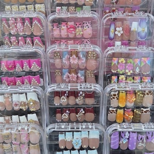 May include: A collection of multiple clear plastic cases containing sets of press-on artificial nails. The nails feature various designs including floral patterns, rhinestones, animal prints, pearls, and colorful geometric shapes in diverse finishes.
