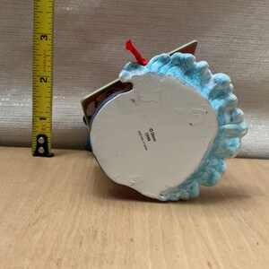 May include: A Disney-branded ornament with a blue, textured base and a red ribbon. The ornament is approximately 3 inches tall. The bottom of the ornament is white with the Disney logo and "China" printed on it.