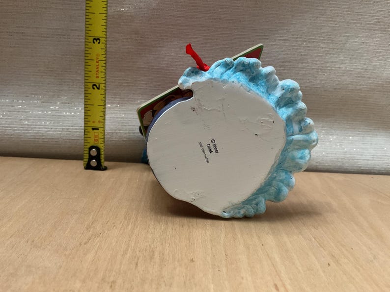 May include: A Disney-branded ornament with a blue, textured base and a red ribbon. The ornament is approximately 3 inches tall. The bottom of the ornament is white with the Disney logo and "China" printed on it.