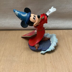 May include: A figurine of Mickey Mouse dressed as a sorcerer, wearing a blue hat with silver stars and a red robe. The figure is posed with one arm raised, wearing white gloves. The base is a dark blue with a light blue wave design.