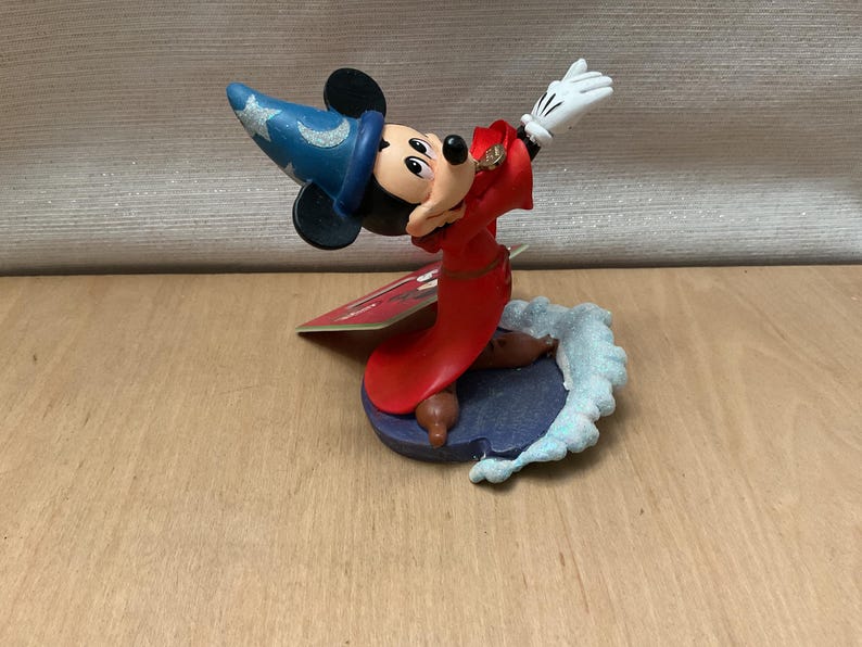 May include: A figurine of Mickey Mouse dressed as a sorcerer, wearing a blue hat with silver stars and a red robe. The figure is posed with one arm raised, wearing white gloves. The base is a dark blue with a light blue wave design.