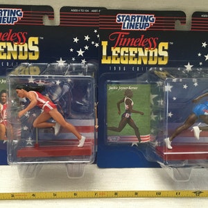May include: Two plastic action figures of female athletes in running poses. The figures are packaged in clear plastic boxes with a red, white, and blue background. The boxes have the text "Starting Lineup Timeless Legends 1996 Edition" on them. The figures are Florence Griffith Joy and Jackie Joyner Kersee.