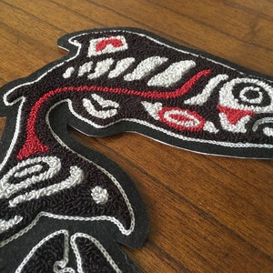 May include: Two embroidered black fabric patches depicting stylized salmon in red, white, and black. The patches are on a wooden surface.