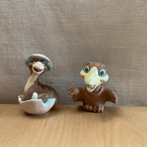 May include: Two brown dinosaur figurines. The left figure is a long-necked creature emerging from a cracked eggshell with a light blue interior. The right figure is a winged creature with a cream-colored beak and large teal eyes, standing upright.