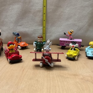 May include: A collection of seven Looney Tunes character plastic toy vehicles, including a red train, orange car, green tractor, red airplane, purple carriage, yellow car, and light blue car, next to a yellow ruler measuring 9 inches tall.