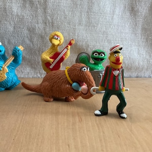 May include: A collection of Sesame Street character figurines, including Cookie Monster, Big Bird, Oscar the Grouch, Bert, and a brown furry creature. Each character is depicted in a playful pose, with some holding musical instruments.