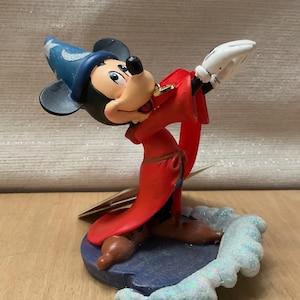 May include: A figurine of Mickey Mouse dressed as a sorcerer, wearing a blue hat with stars and a red robe. The figure is posed with arms outstretched, standing on a blue base with a glittery, icy design.