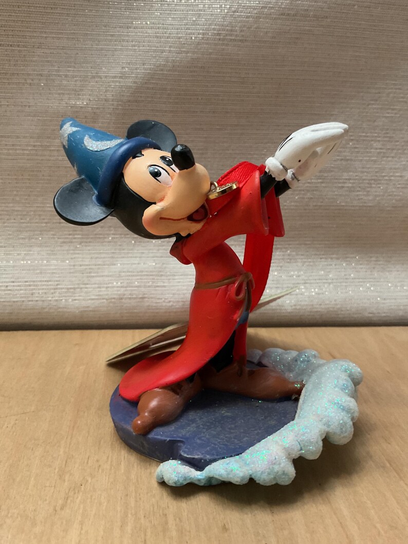 May include: A figurine of Mickey Mouse dressed as a sorcerer, wearing a blue hat with stars and a red robe. The figure is posed with arms outstretched, standing on a blue base with a glittery, icy design.