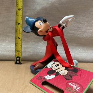 May include: A Mickey Mouse figurine in a red robe and blue hat, with a gold necklace. The figurine is approximately 4 inches tall. A red and green Disney Store tag is attached, featuring Mickey Mouse in a holiday design.