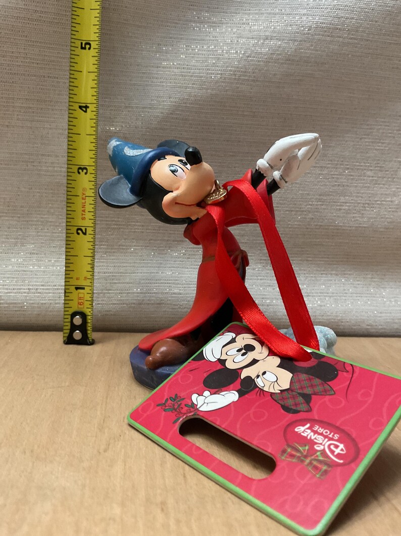 May include: A Mickey Mouse figurine in a red robe and blue hat, with a gold necklace. The figurine is approximately 4 inches tall. A red and green Disney Store tag is attached, featuring Mickey Mouse in a holiday design.
