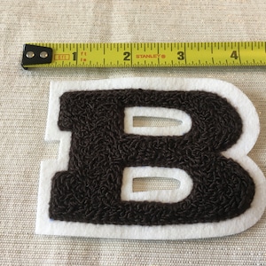 May include: A brown, embroidered letter "B" patch with a white border. The patch is approximately 3 inches wide. A yellow and black measuring tape is placed above the patch.