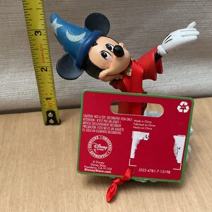 May include: A Mickey Mouse figurine dressed as a sorcerer, wearing a blue hat with stars and a red robe. The figurine is approximately 3 inches tall. A red tag with text is attached.