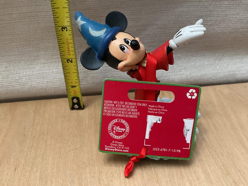 May include: A Mickey Mouse figurine dressed as a sorcerer, wearing a blue hat with stars and a red robe. The figurine is approximately 3 inches tall. A red tag with text is attached.