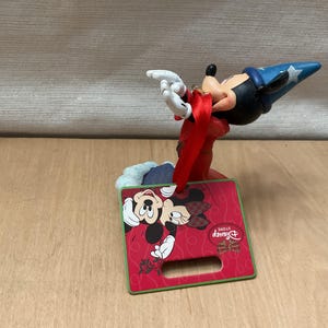 May include: A Mickey Mouse figurine in a sorcerer's hat, with a red and green Christmas-themed display. The display features Mickey and Minnie Mouse, with the Disney Store logo. The figurine is on a wooden surface.
