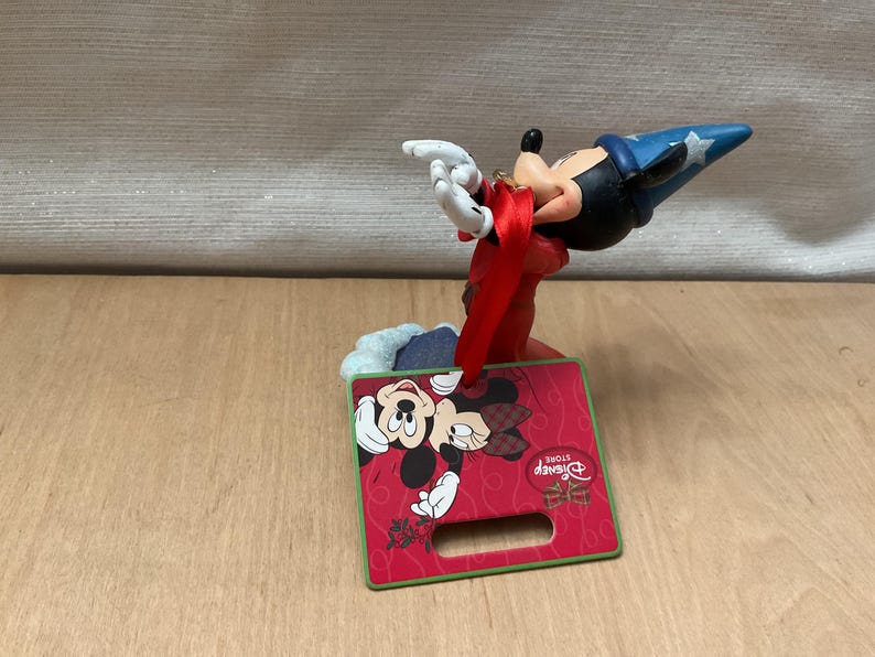 May include: A Mickey Mouse figurine in a sorcerer's hat, with a red and green Christmas-themed display. The display features Mickey and Minnie Mouse, with the Disney Store logo. The figurine is on a wooden surface.