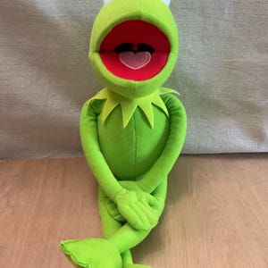 May include: A bright green plush frog doll with a wide open mouth, a red interior, and a pink tongue. The character features large white eyes with black pupils, a pointed collar around the neck, and long, slender arms and legs in a seated position.