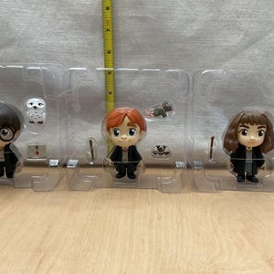 May include: A set of three stylized figurines of characters from a fantasy series. The figures are in a clear plastic tray. The characters include a boy with glasses, a boy with red hair, and a girl with brown hair. Each figure is approximately 3 inches tall.