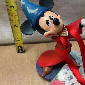 May include: A Mickey Mouse figurine dressed as a sorcerer, wearing a blue hat and red robe. The figurine has a gold charm that says "2013". A yellow tape measure is next to the figurine.