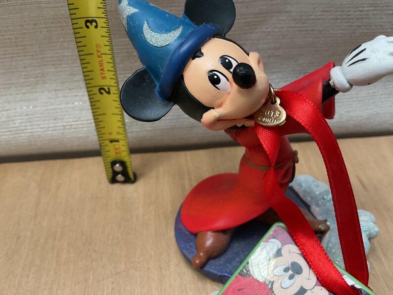 May include: A Mickey Mouse figurine dressed as a sorcerer, wearing a blue hat and red robe. The figurine has a gold charm that says "2013". A yellow tape measure is next to the figurine.