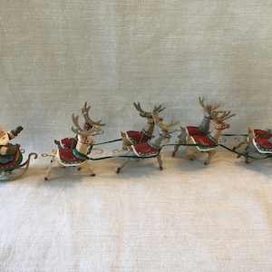 May include: A decorative Santa Claus figurine sitting in a green and red sleigh, pulled by a team of five reindeer. Each reindeer wears a red blanket with a name embroidered in gold, connected by thin green cords to the sleigh and each other.