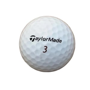 May include: A white golf ball with a dimpled surface featuring the black TaylorMade logo and the number 3 printed in black. The spherical object has a textured exterior designed for aerodynamic performance during flight on the golf course.