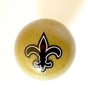 May include: A spherical object with a gold glitter finish featuring a black fleur-de-lis emblem centered on the front. The emblem is outlined in white, creating a high-contrast design against the shimmering, textured gold surface of the sphere.