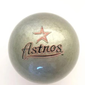 May include: A spherical, metallic silver object featuring the Houston Astros logo in the center. The logo consists of a red-outlined star positioned above the word Astros written in a black, cursive script with a thin red outline and a trademark symbol.