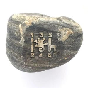 May include: A smooth, grey, oval-shaped river stone featuring an engraved manual transmission gear shift pattern. The pattern includes the numbers 1, 2, 3, 4, 5, 6, and R, with a small lizard silhouette positioned in the center of the gear layout.