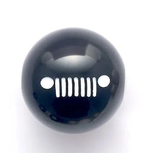 May include: A spherical black gear shift knob featuring a white graphic of a seven-slot grille flanked by two circular headlights, mimicking the iconic front end design of a vehicle. The surface is smooth, glossy, and reflective.